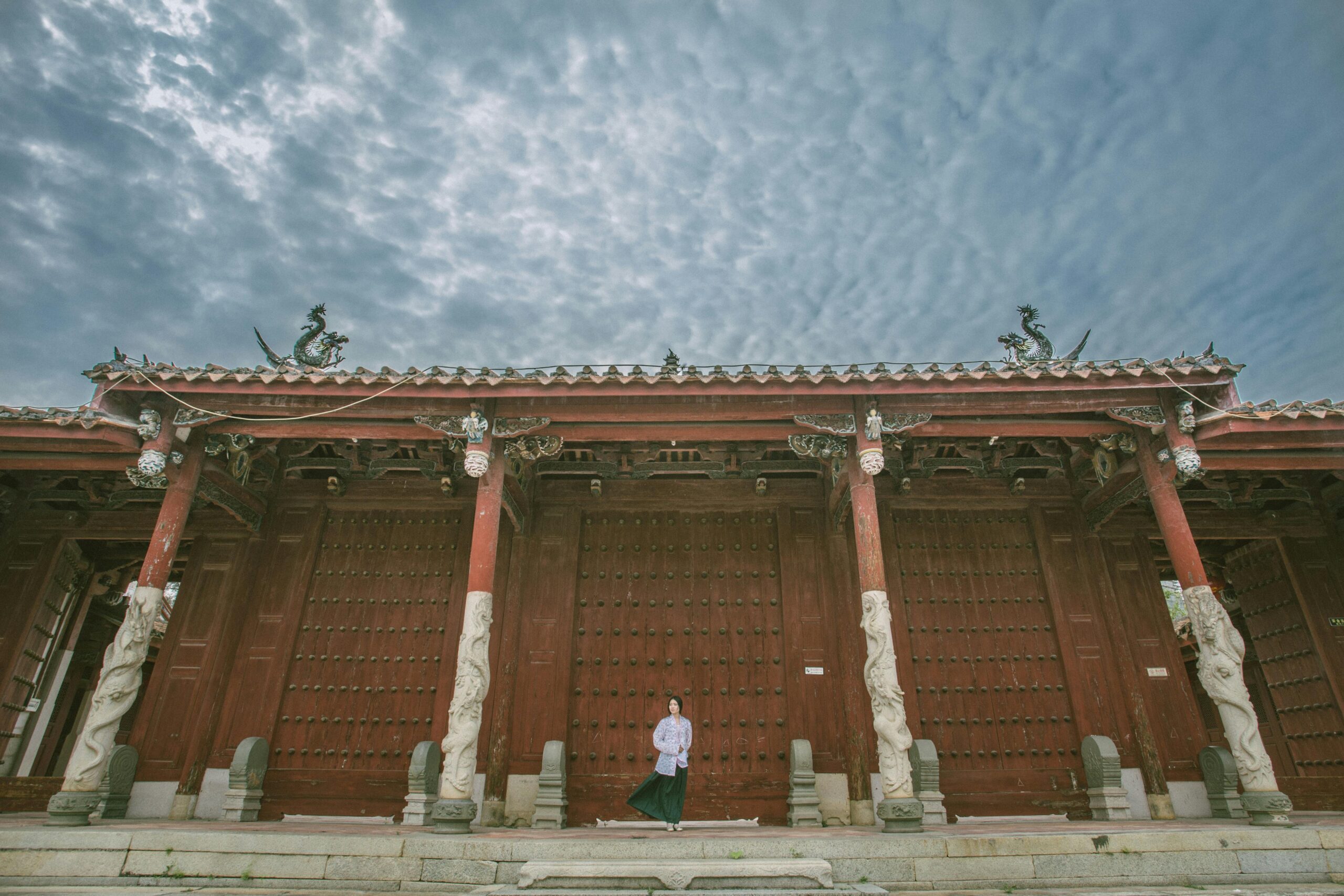 An adult woman stands in front of a historic Chinese temple gate with ornate dragon pillars under an overcast sky.